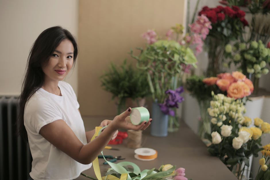 A smiling young woman with long dark hair, wearing a white t-shirt, holds a roll of light green floral wrapping ribbon in her right hand and a pair of scissors in her left hand, standing at a workspac