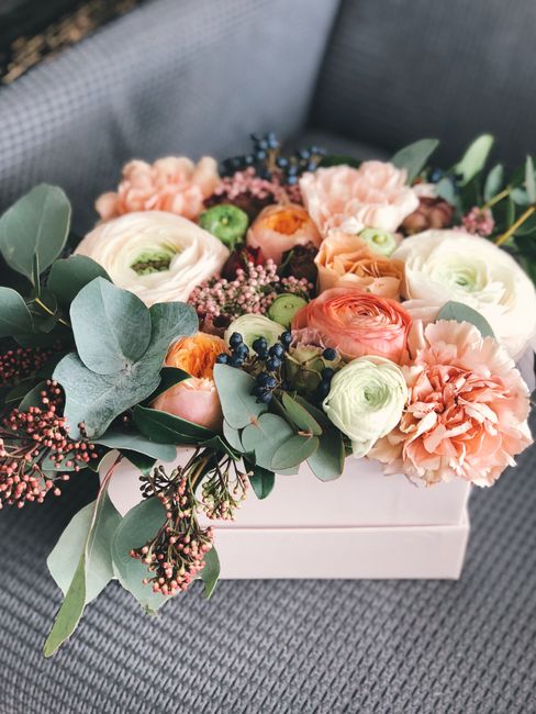 A floral bouquet arranged in a white rectangular box featuring pastel-colored ranunculus, carnations, and roses in shades of peach, white, and soft pink, accented with sprigs of pink baby's breath, da