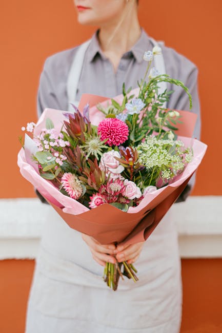 A person wearing a light grey shirt and white apron is holding a vibrant bouquet of fresh flowers, wrapped in soft pink and peach-colored paper. The bouquet features an array of pink, white, and green