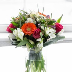 Bathed in the gentle daylight characteristic of a Surrey morning, this bouquet reflects both local charm and sophisticated floristry. The arrangement draws immediate attention to a distinctive orange Gerbera daisy, luminous and cheerful-a floral sunburst harmonising with jewel-toned roses in shades of deep fuchsia and crimson, each layered petal unfolding with a soft promise of romance. White alstroemeria lilies, recognisable by their slender, striped petals and delicate form, weave through the composition, reminiscent of the pristine gardens bordering Surrey's leafy avenues. Scattered among these, clusters of tiny white chrysanthemums bring a subtle frothiness, their light, lacy texture playing against the more substantial blooms. Creamy rosebuds and pale yellow roses contribute a whisper of softness, while splashes of lavender and muted purple add gentle dimension, echoing the tranquil borders of local woodland walks. Broad, glossy leaves-perhaps inspired by the lush foliage found in RHS Garden Wisley-wrap gracefully around the base, while spiky greens introduce movement and structure. The entire arrangement is anchored in a pure, cylindrical glass vase, fresh stems glowing in pristine water. This bouquet conjures memories of wandering flower markets in Guildford or sending a bright gesture to a friend in Woking: whether as a gift or a centrepiece, it radiates comfort, delight, and the thoughtful artistry of a seasoned Surrey florist.